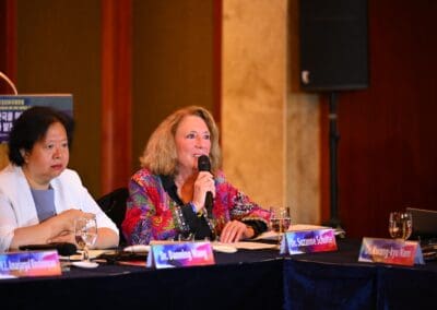 Two women sit at a conference table during the International Forum. One woman speaks into a microphone, while the other listens. Nameplates, glasses of water, and microphones are visible on the table at the One Korea 2025 event.