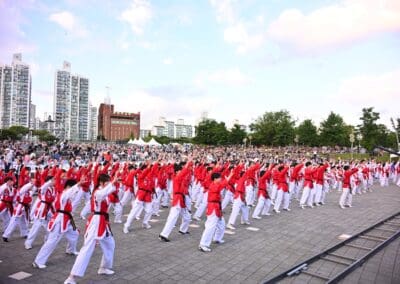A large group of people in red and white martial arts uniforms performs synchronized moves outdoors at Hangang Festa 2025, with a crowd watching and tall buildings in the background, celebrating the Korean Dream.