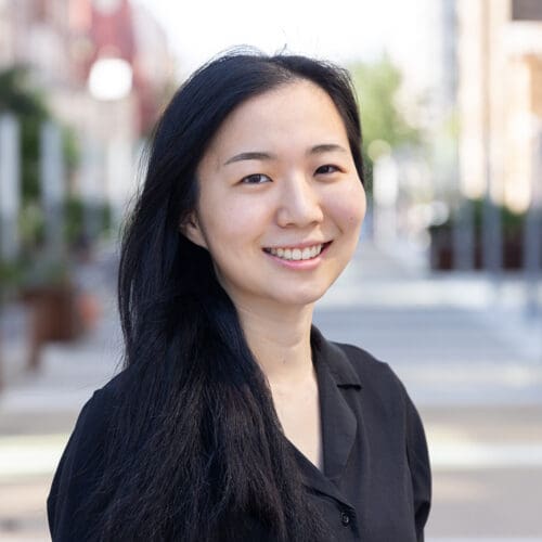 Mrs. Takae Goto, a woman with long black hair wearing a black shirt, stands outdoors on a city street, smiling at the camera with buildings and trees blurred in the background.