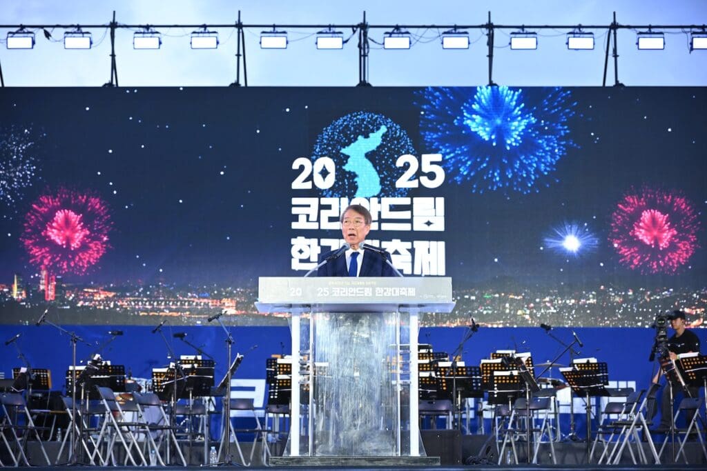 A man speaks at a podium on stage with an orchestra behind him and a large screen displaying 