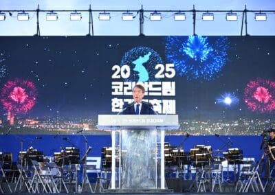 A man speaks at a podium on stage with an orchestra behind him and a large screen displaying "2025" and fireworks graphics, celebrating the spirit of the Korean Dream at Hangang Festa.