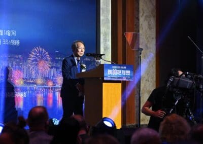 A man in a suit stands at a podium speaking at the International Forum on One Korea, with cameras and an audience present; a cityscape and fireworks are displayed on a screen behind him.