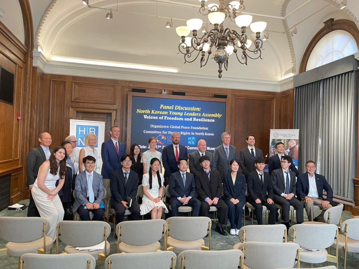 A group of people in formal attire pose for a photo in a conference room after the North Korean Young Leaders Assembly panel discussion, with banners and empty chairs visible in the background.