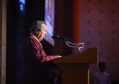 A man in a maroon suit speaks at a podium with a microphone and a glass of water, addressing the audience at the International Forum on One Korea in a dimly lit indoor setting.