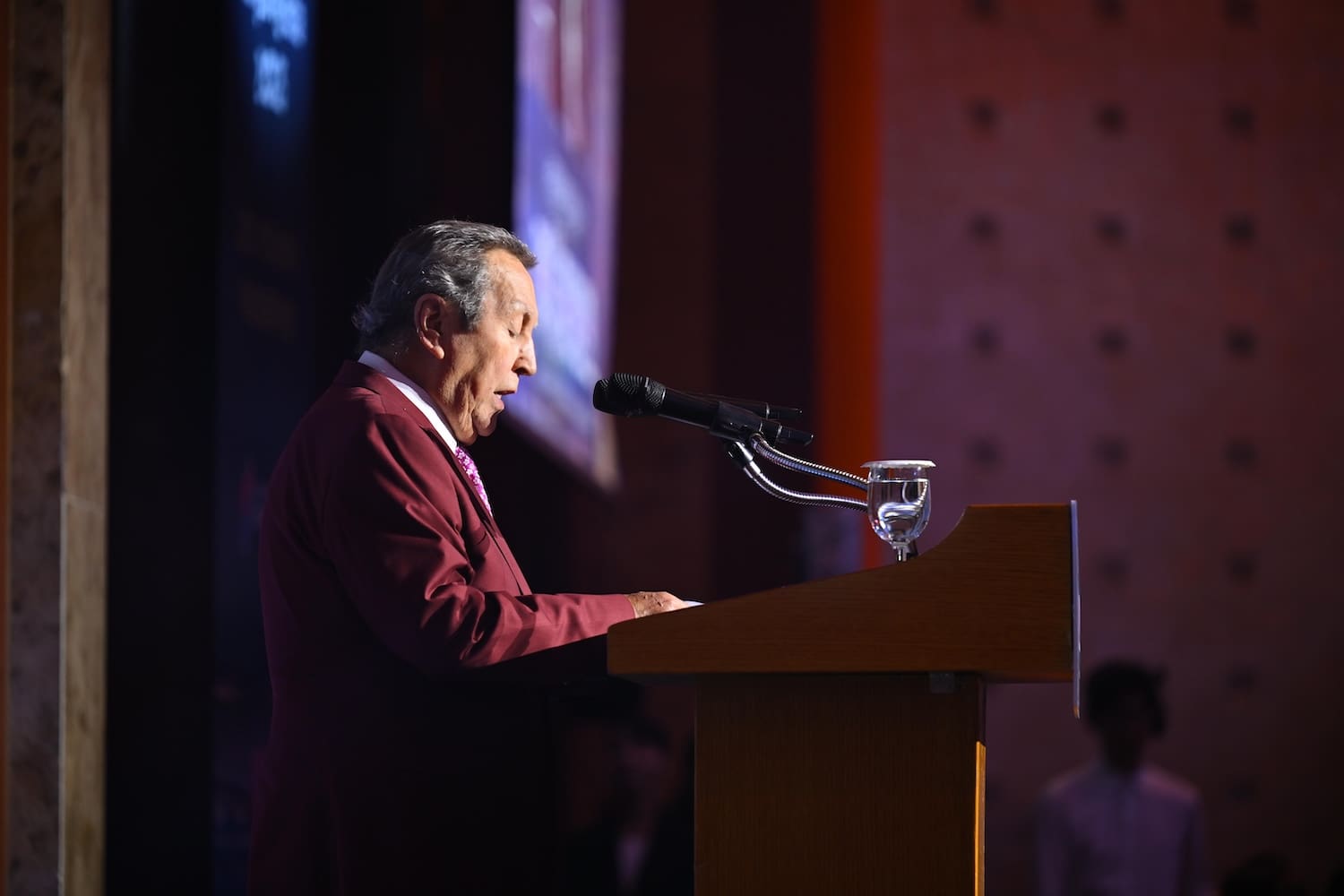 A man in a maroon suit speaks at a podium with a microphone and a glass of water, addressing the audience at the International Forum on One Korea in a dimly lit indoor setting.