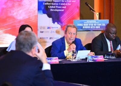 A man in a blue blazer speaks into a microphone at the International Forum on One Korea, seated at a conference table with other attendees and a colorful banner in the background.