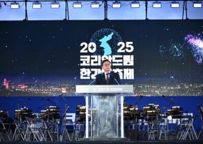 A man in a suit speaks at a podium on stage with an orchestra behind him; a screen displays "2025" and Korean text, highlighting the Korean Dream during Hangang Festa, with fireworks and a map graphic in the background.