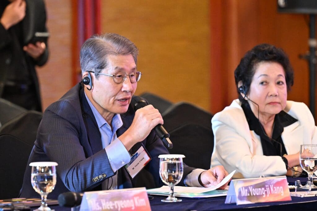 At an International Forum on One Korea 2025, two people sit at a conference table with microphones and earpieces; the man on the left is speaking, while the woman on the right listens. Nameplates and water glasses are visible.