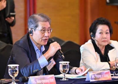 At an International Forum on One Korea 2025, two people sit at a conference table with microphones and earpieces; the man on the left is speaking, while the woman on the right listens. Nameplates and water glasses are visible.