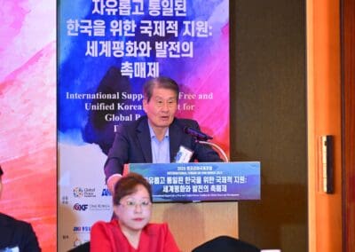 A man in a suit speaks at a podium during the International Forum on One Korea, with a blue and pink bilingual banner in Korean and English displayed behind him.