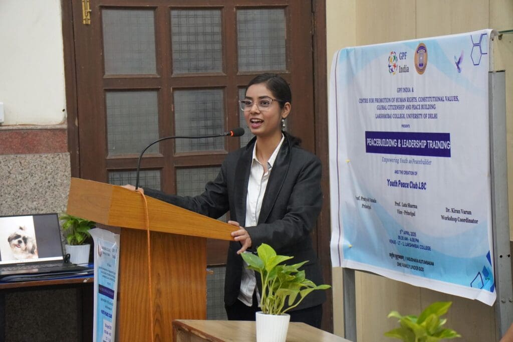 A woman in a black blazer speaks at a wooden podium during a leadership training event, inspiring others to Lead with Purpose. A banner and potted plants are visible in the background.