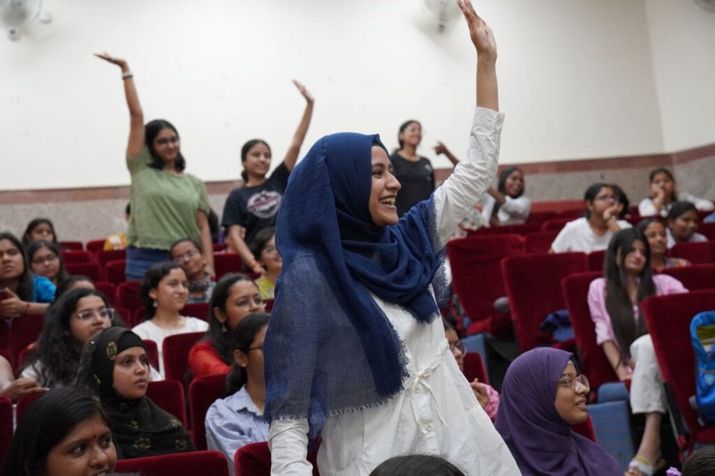 A group of women and girls sit in red auditorium seats, several raising their hands and smiling as they participate in a GPF India event focused on youth empowerment.