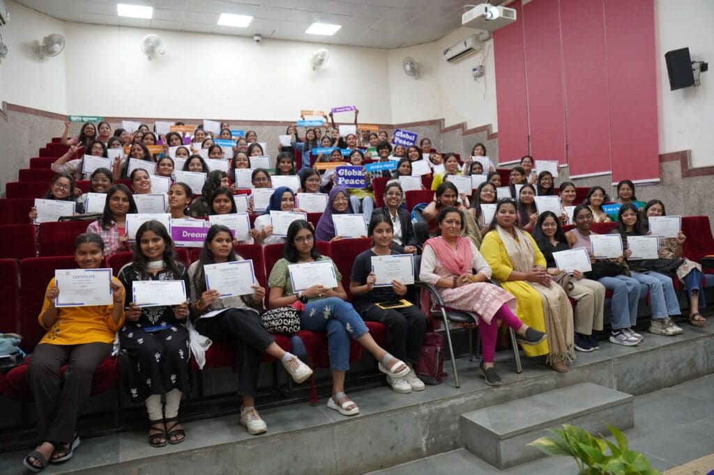 A large group of women and girls sit in an auditorium, holding certificates and smiling at the camera; some hold signs with positive messages, celebrating youth leadership at a GPF India event.