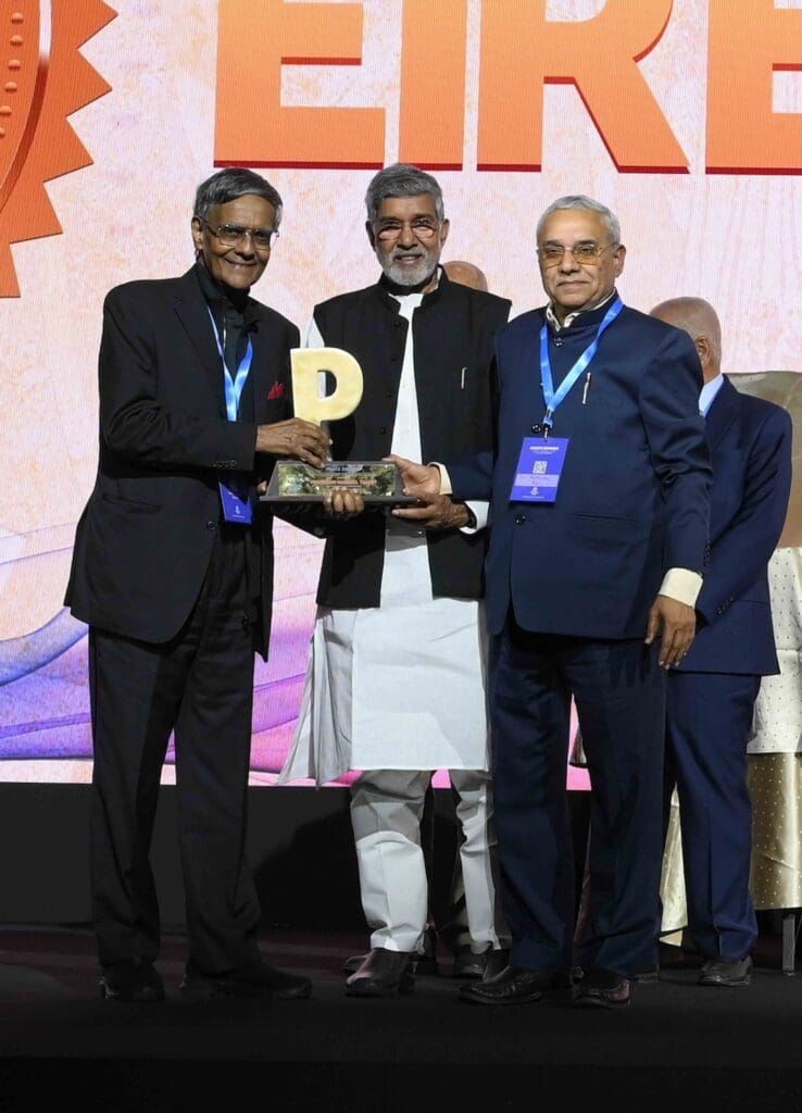 Three men in formal attire stand on stage at the Global Justice Summit; one holds a trophy, while the others pose beside him in front of a large, colorful backdrop featuring Dr. Markandey Rai.