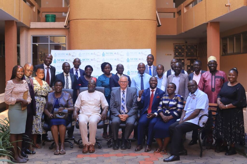 A group of men and women pose for a formal photo outside a building in front of a banner, with some seated and others standing behind them at the Uganda Transforming Education Workshop August 2025.