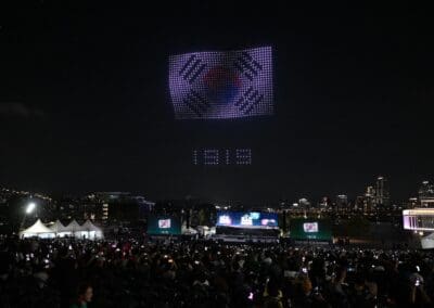 A large crowd watches a mesmerizing drone show at night, with drones forming the South Korean flag and numbers in the sky above a cityscape.