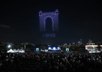 A nighttime outdoor drone show draws a large crowd as drones form an illuminated arch and letters in the sky above a sparkling cityscape.