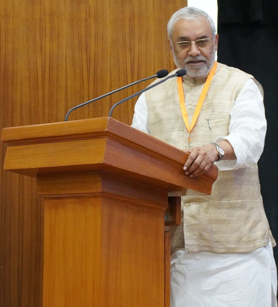 A man in a beige vest and white kurta stands at a wooden podium with microphones, addressing an indoor event at IIT Delhi focused on building a sustainable future.