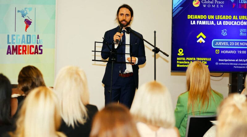 A man in a suit speaks at a podium to an audience. Behind him, a screen displays event information and a banner reading