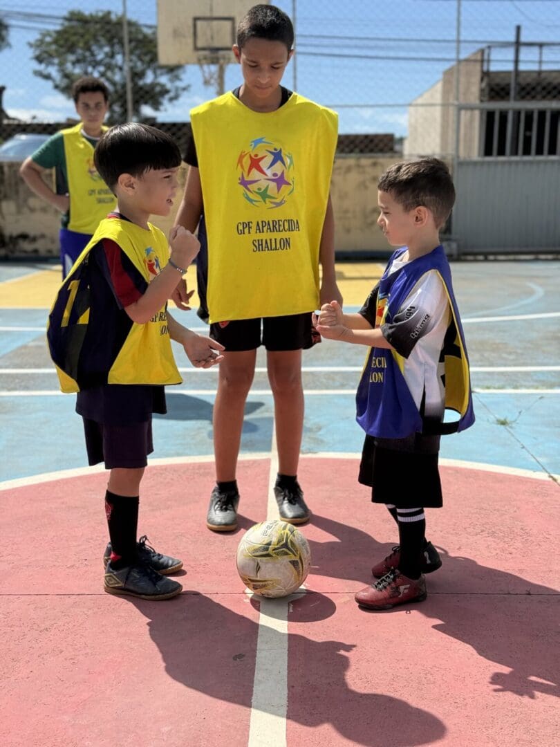 Two young boys in sports uniforms play rock-paper-scissors at the center of an outdoor basketball court, while an older boy—illustrating youth leadership development—stands behind them. A soccer ball is placed between them.