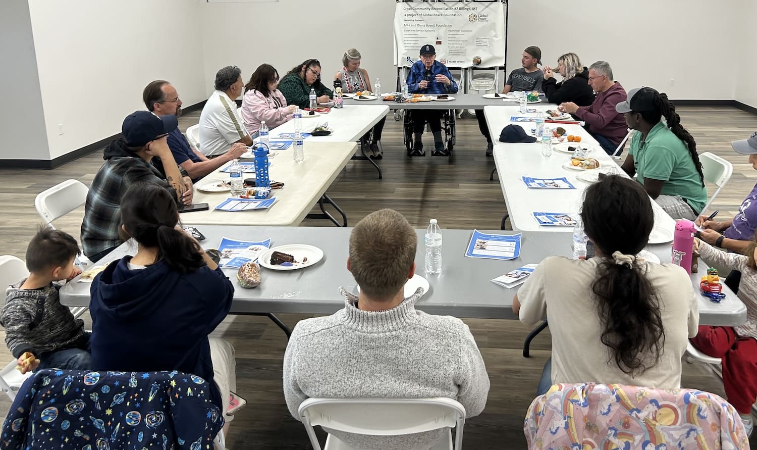 A diverse community sits around tables in a meeting room, listening to a speaker at the front; papers, food, and drinks are on the tables during the reconciliation project.