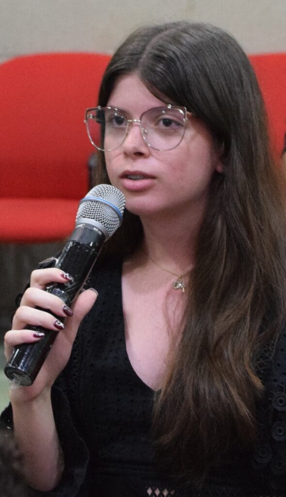 A young woman with long brown hair and glasses speaks into a microphone about Korean Peninsula Unification. She wears a black top and sits in front of red chairs.