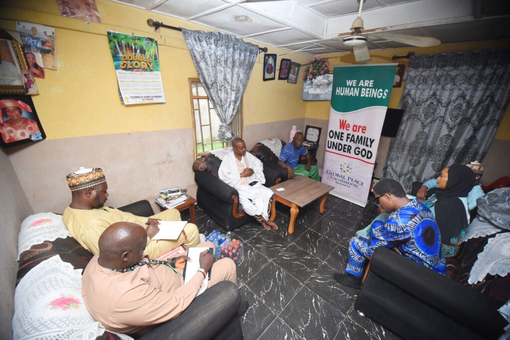 A group of people sit in a living room having a discussion about peaceful coexistence; posters on the wall include messages about unity and faith.