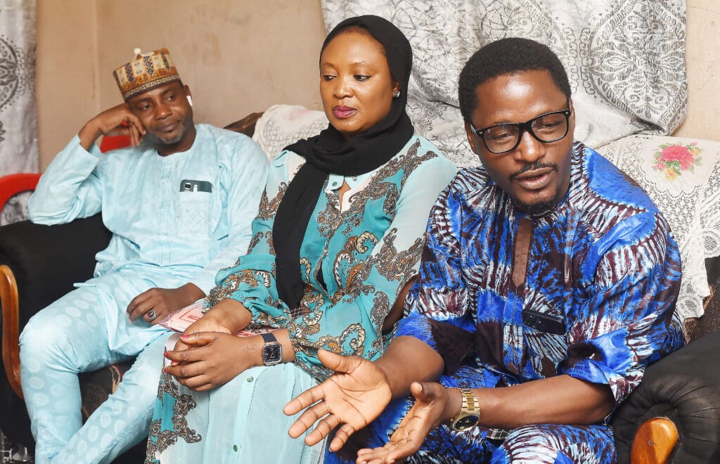 Three people sit on a couch indoors, having a conversation about peaceful coexistence. The man on the right gestures with his hands while the others listen. They wear patterned traditional clothing.