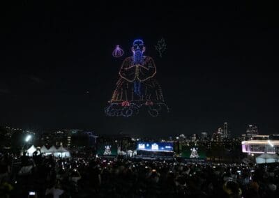 A crowd watches a nighttime drone show as lights form an image of a seated figure with a staff and leaf above the cityscape.