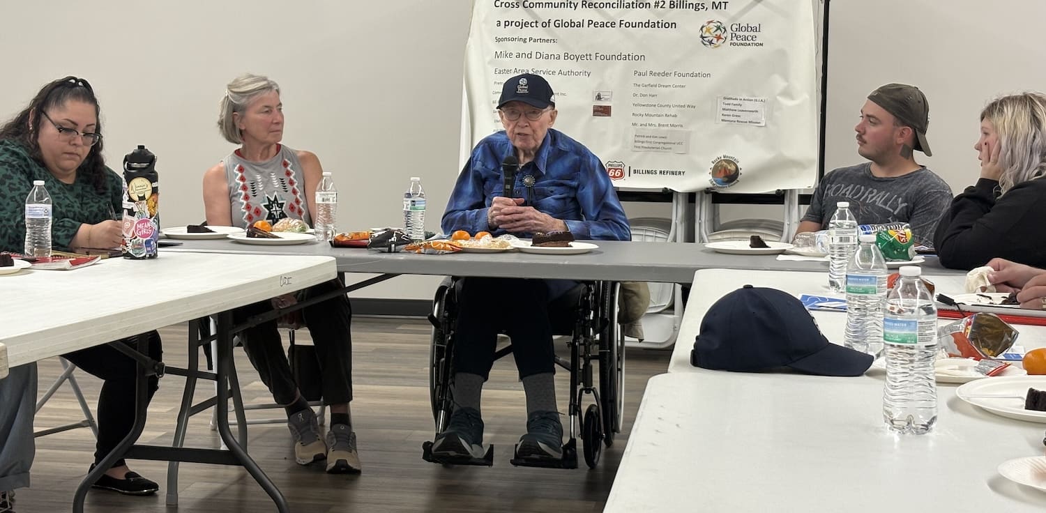 A group of people sit around tables with food and drinks, listening to Dr. Don Harr, an elderly man in a wheelchair, speak about community reconciliation at a Montana event. A poster board is displayed behind him.