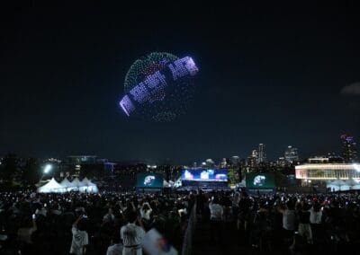 A large crowd watches a spectacular nighttime drone show in the sky above a city, with illuminated buildings in the background and many people taking photos.