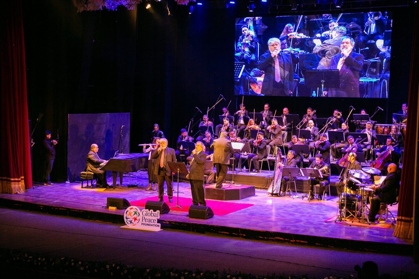 A large orchestra performs on stage with three male vocalists in front, singing in Guaraní language; a screen above displays a close-up of the singers. The stage features a