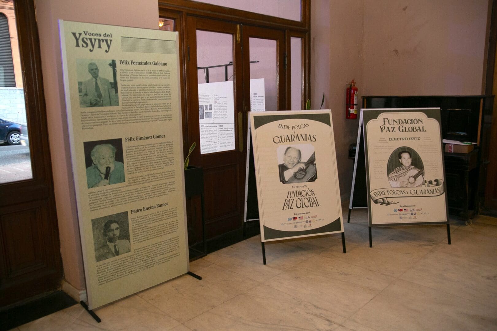 Three informational display boards stand in a hallway, featuring text and black-and-white photos about individuals and a foundation, highlighting aspects of Paraguayan cultural heritage, with wooden doors and white walls in the background.