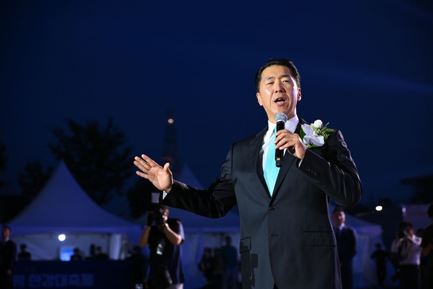 A man in a suit with a flower boutonnière speaks into a microphone on an outdoor stage at night, with tents and people—many from Gen Z—gathered for a Global Peace Foundation event promoting Korea unification in the background.