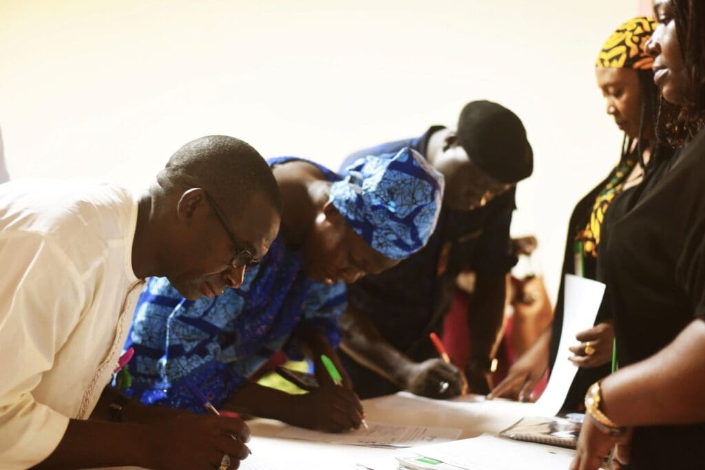 Several people stand at a table, signing papers or documents—some holding pens and wearing brightly colored clothing—demonstrating their commitment to youth leadership and sustainable development.