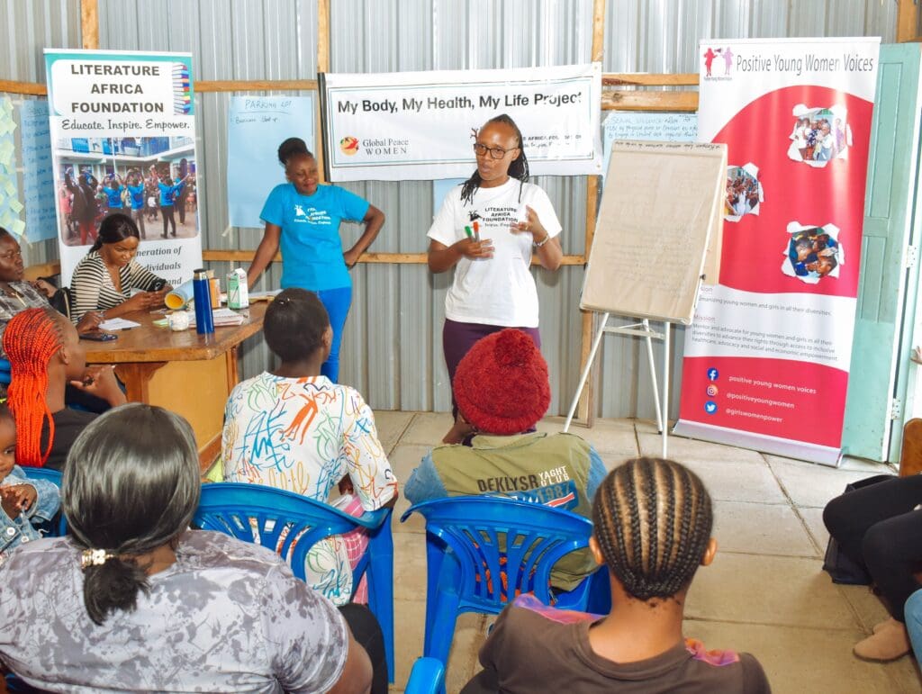 A woman demonstrates leadership as she stands and speaks to a seated group in a room with banners for youth and health organizations displayed on the walls, highlighting women empowerment.