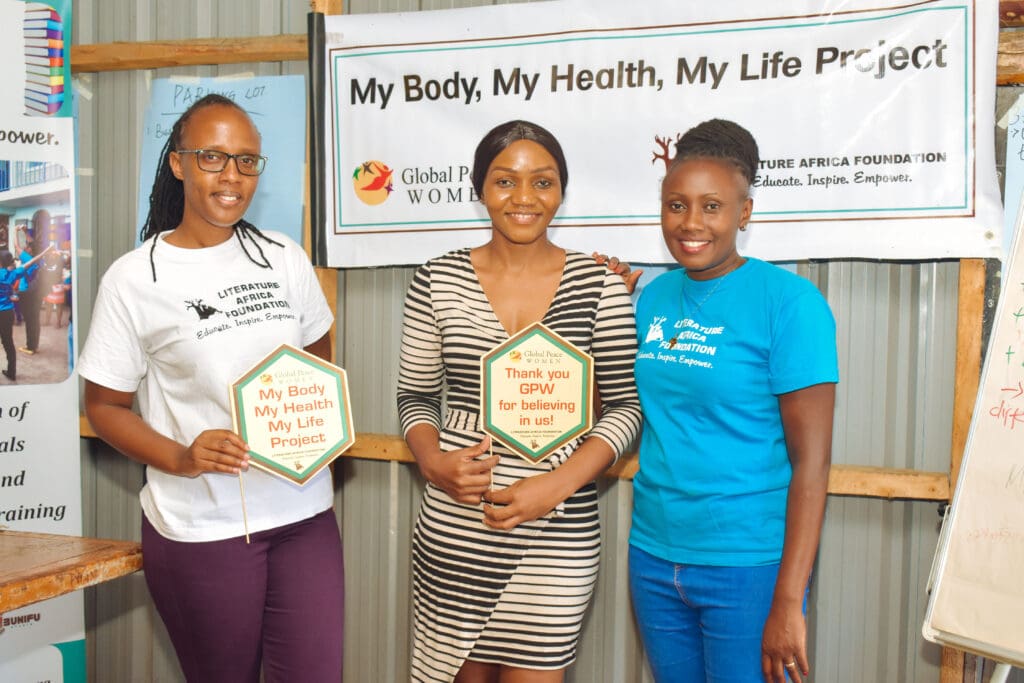 Three women stand indoors holding signs promoting the 