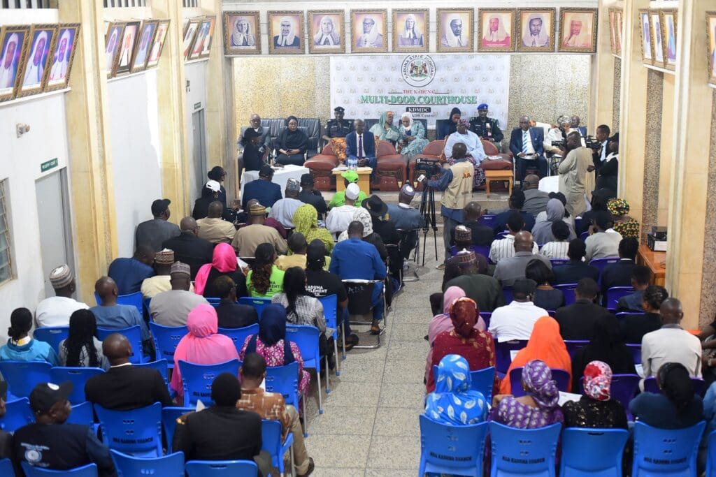 A large group of Peacebuilders seated in rows face a panel at the front of a conference room decorated with portraits and an official Kaduna Settlement Week banner organized by GPF Nigeria.