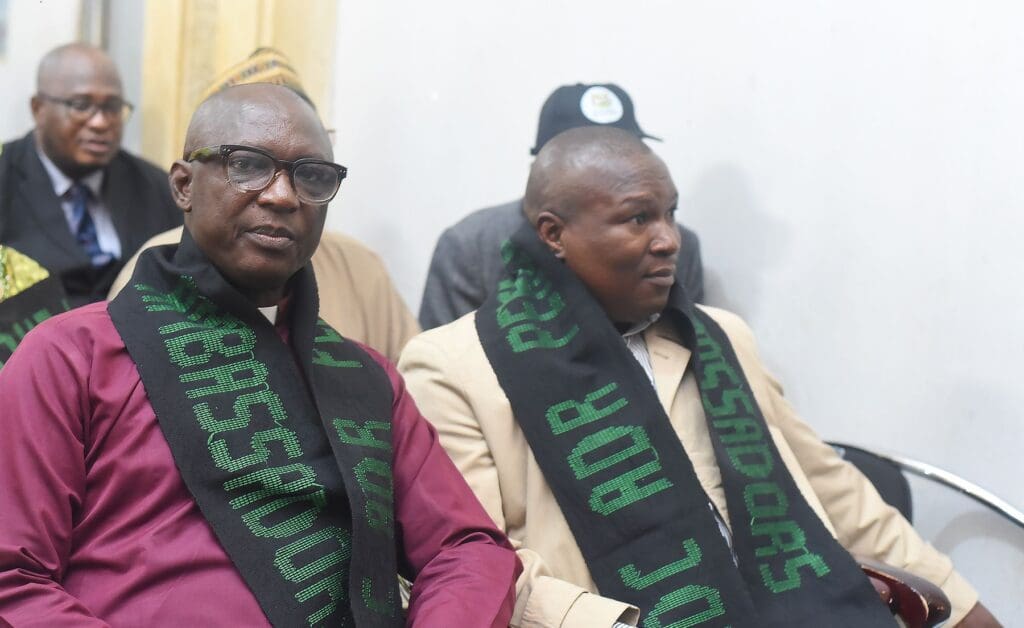 Two men sit side by side wearing dark scarves with green lettering at an indoor event during Kaduna Settlement Week; other Peacebuilders and GPF Nigeria attendees are visible in the background.