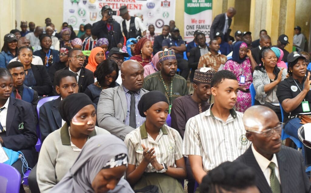 A diverse group of students and adults, some in uniforms, sit in rows at an indoor event for Kaduna Settlement Week by GPF Nigeria, facing forward with a banner in the background—a gathering of aspiring Peacebuilders.