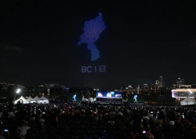 A nighttime crowd enjoys a dazzling drone show as hundreds of lights form the Korean Peninsula map and the text "DCIE" in the sky above the cityscape.