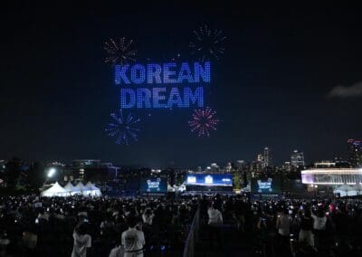 A large crowd watches a dazzling drone show in the night sky, where drones spell out "KOREAN DREAM" and fireworks light up the cityscape near a river.