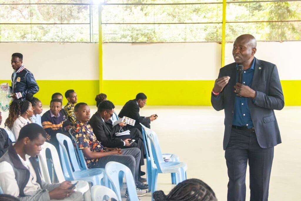 A man in a suit speaks into a microphone to a seated audience of young adult changemakers in a bright indoor space.