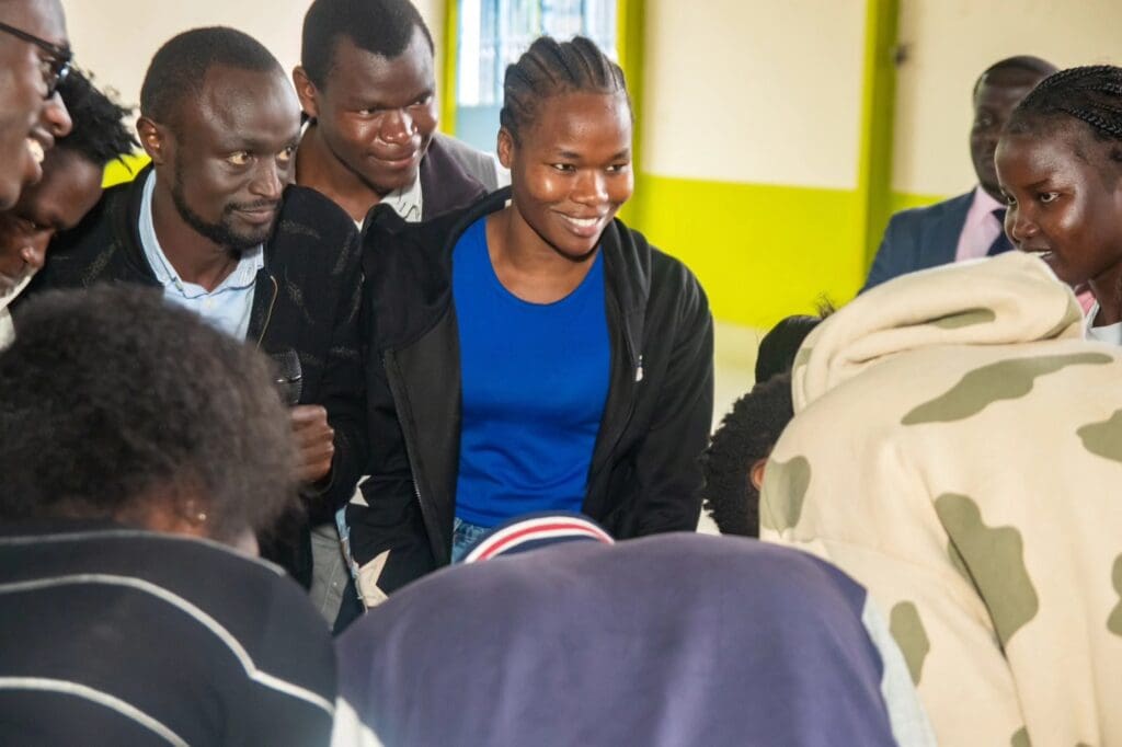 A group of young adults, some smiling, gather closely indoors around a table or object, with bright yellow-green walls in the background—embodying the spirit of changemakers united for education and peace.