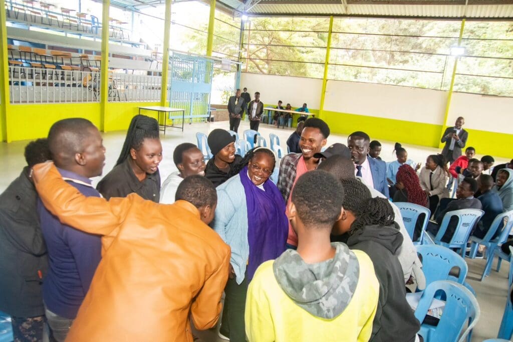 A group of changemakers stands and interacts in a brightly lit indoor hall with rows of blue chairs and a few people seated in the background.