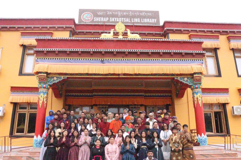A large group of people pose in front of the Sherab Skatdsel Ling Library in Leh, its traditional architecture and signage above the entrance setting the scene for a memorable moment at the World Peace Conference.