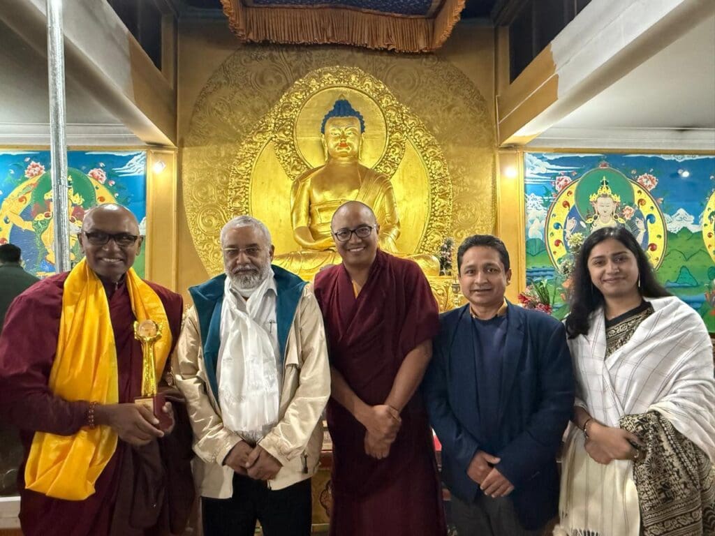 Five people stand indoors in front of a large golden Buddha statue, with colorful wall art on both sides. The group, gathered for the World Peace Conference in Leh, includes two monks in robes and three others in formal attire.