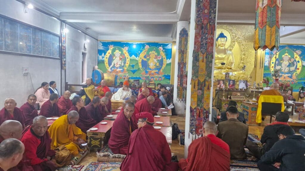 A group of monks and people sit on the floor in a decorated temple in Leh during the World Peace Conference, with a large Buddha statue and colorful wall art in the background.