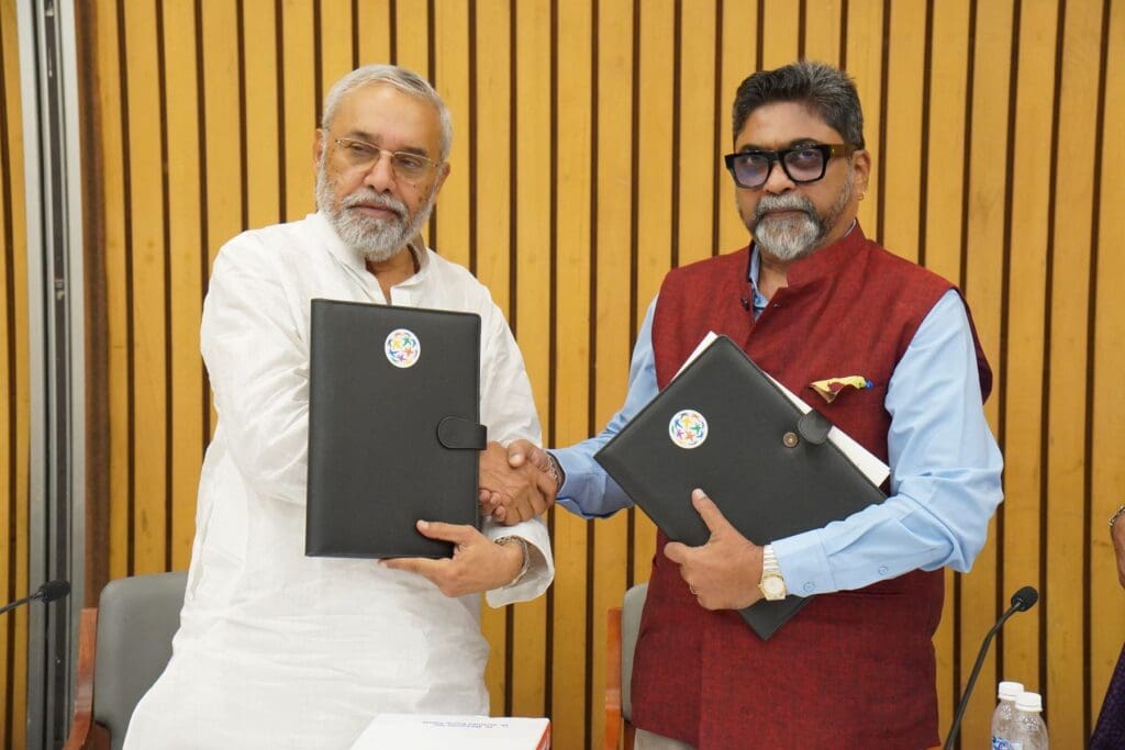 Two men in formal attire shake hands while holding black folders, standing in front of a wooden panel wall at an indoor event organized by ASWWF and GPF India to support acid attack survivors.