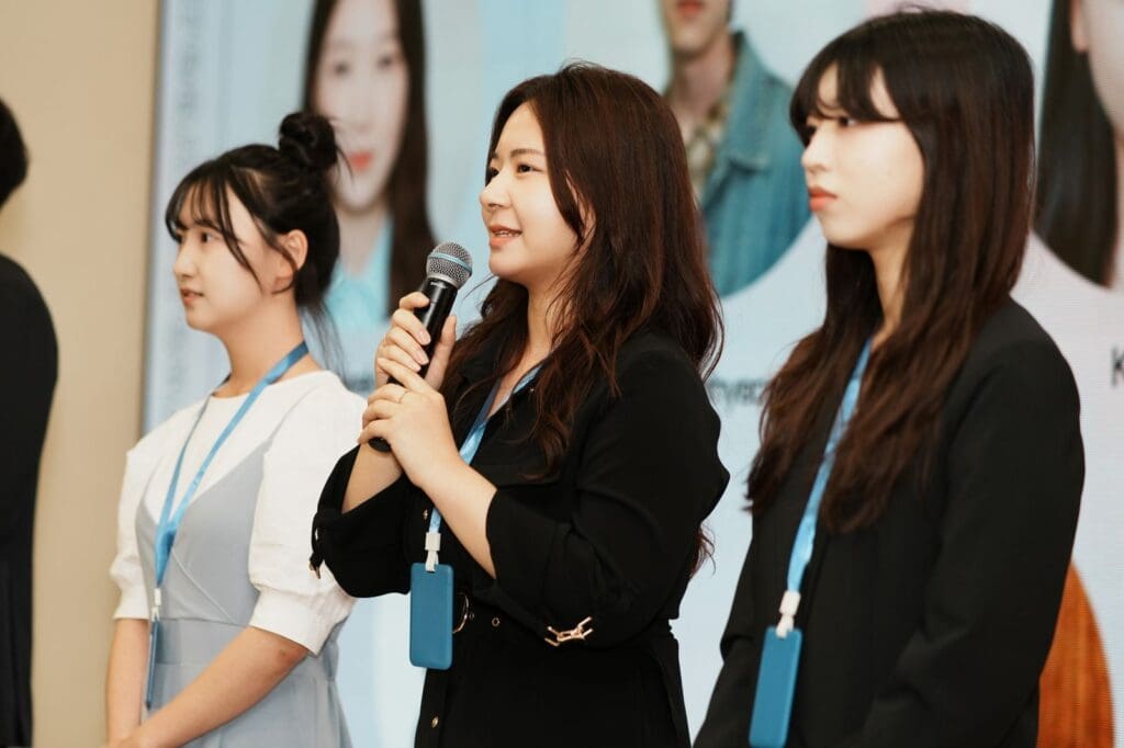 Three women wearing name badges stand in a row; the middle woman speaks into a microphone. A large display with photos and text is visible in the background, highlighting Young Leaders Mongolia and their commitment to empowering youth Mongolia.